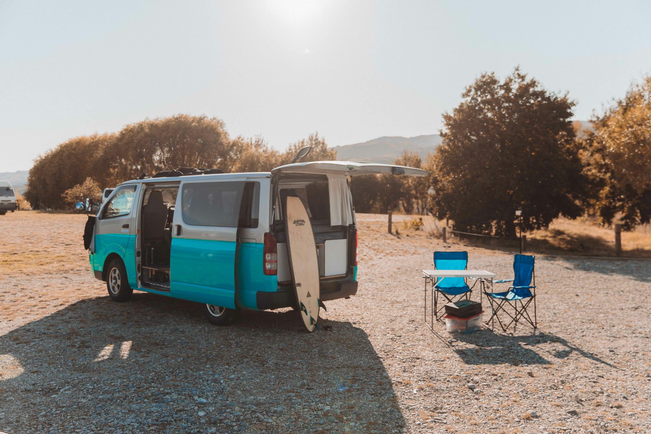 Parked camper in remote area with surfboard, table and chairs.